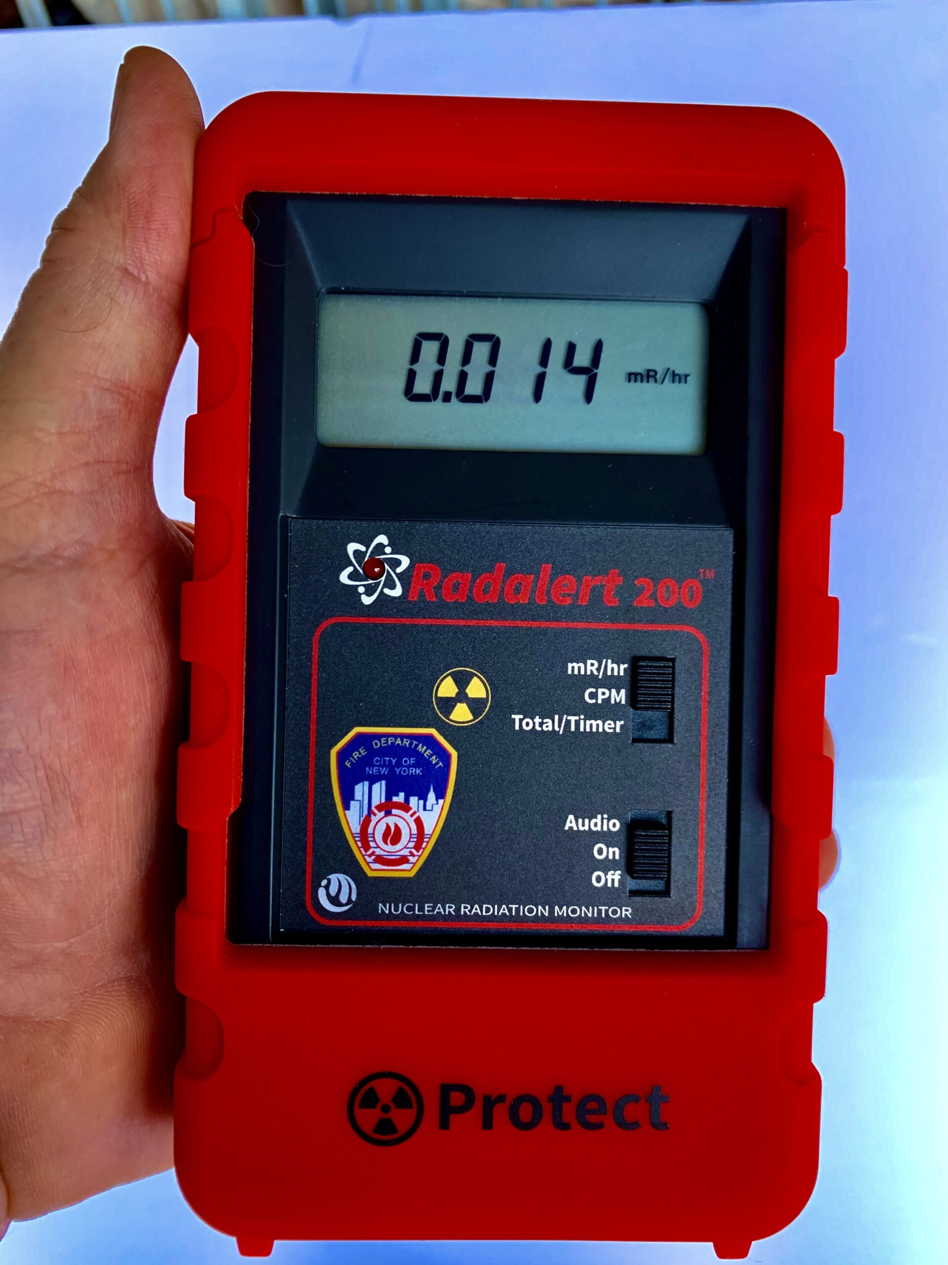 A hand holds a Radalert 200 professional Geiger counter in a red Protect® silicone boot, with large easy to read LCD display. The tactile slide switches are easy to use with safety gloves on, and favored by firefighters and first responders.